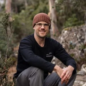 Andy Crawford, a man wearing glasses and a beanie, sits on a rock in the woods.