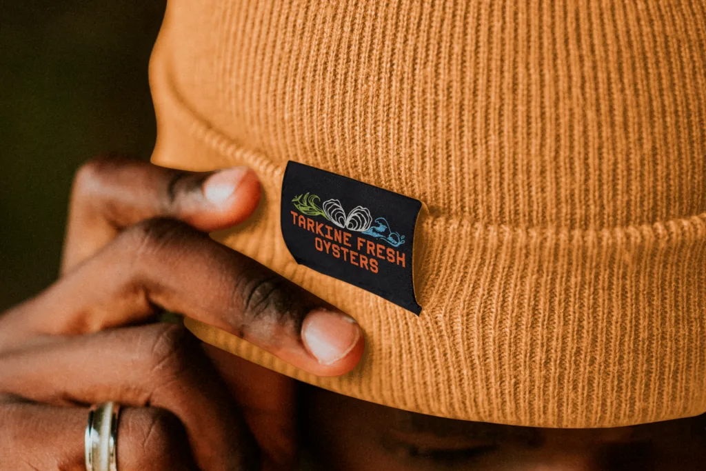 person wearing an orange beanie with a "tarkine fresh oysters" label, touching the edge with fingers.
