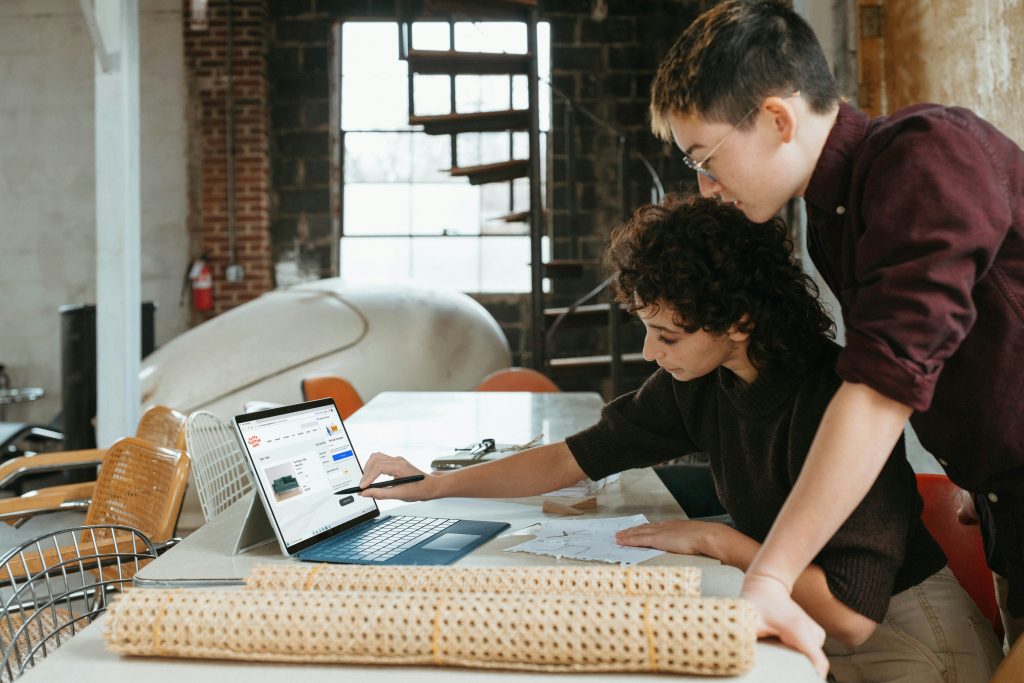 Two people look at a laptop screen together in a modern workspace, with rolled-up materials and papers on the table in front of them, collaborating on strategies for starting a business.