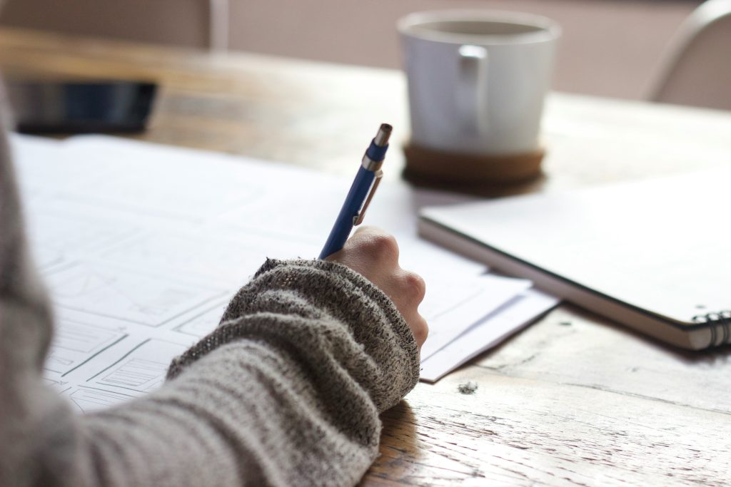 Person wearing a jumper writes on paper at a wooden table with a coffee mug, spiral notebook, and mobile nearby, drawing inspiration for AI copywriting.