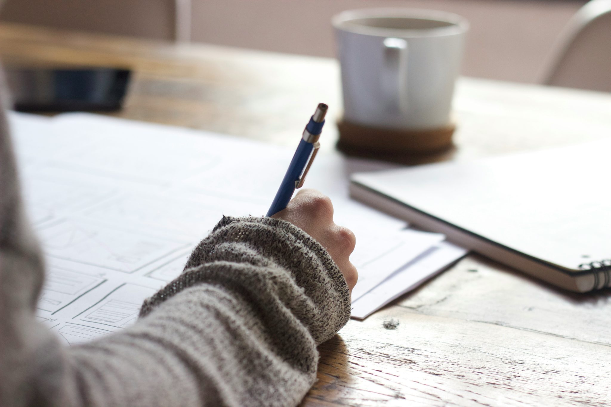Person wearing a jumper writes on paper at a wooden table with a coffee mug, spiral notebook, and mobile nearby, drawing inspiration for AI copywriting.