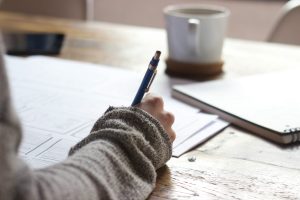 Person wearing a jumper writes on paper at a wooden table with a coffee mug, spiral notebook, and mobile nearby, drawing inspiration for AI copywriting.